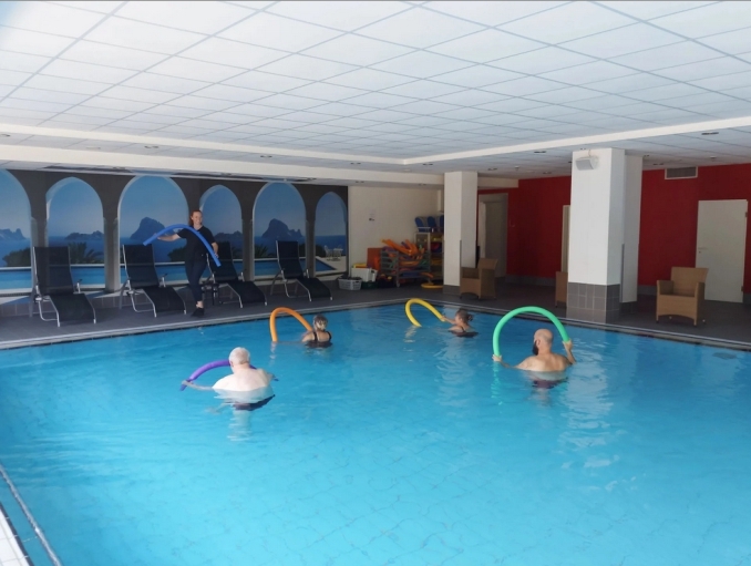 Aqua Fit in unserem Schwimmbad. Die Trainerin steht am Beckenrand und gibt Anweisungen, die Teilnehmer stehen im Wasser und führen die Übungen mit Schwimmnudeln durch.
