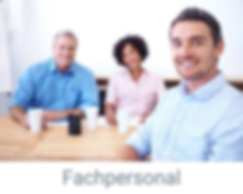 Ein junger Mann sitzt im Vordergrund an einem Tisch, eine Frau und ein Mann sitzen im Hintergrund. Alle lächeln in die Kamera. Das Bild hat den Titel Fachpersonal