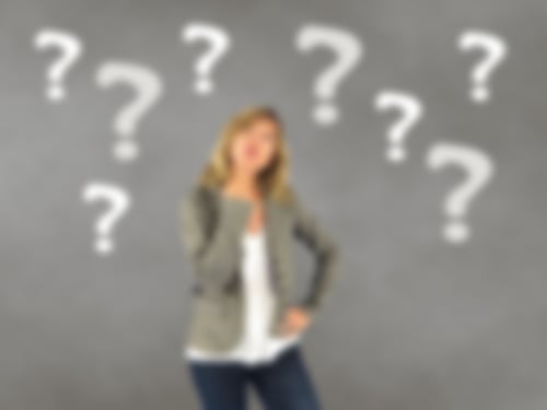 eine Frau mit Jacket und Bluse steht vor einer grauen Wand voller Fragezeichen. Sie schaut nachdenklich und hat die Hand am Kinn