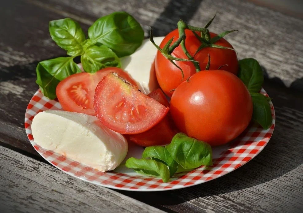 3Tomaten und ein aufgeschnittener Weichkäse liegen auf einem rot-weiß karierten Teller, der mit Basilikumblättern dekoriert ist.