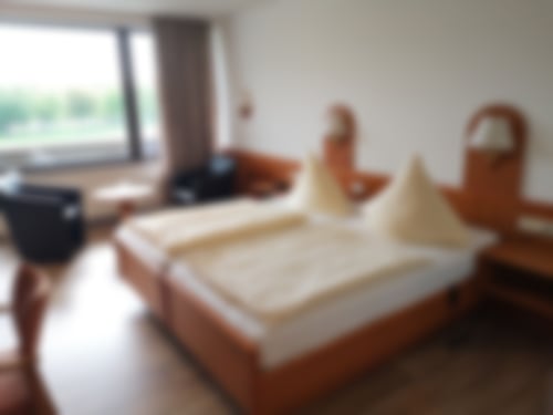 Ein Doppelzimmer mit Doppelbett und Nachttisch, TV, Schreibtisch, Sesseln und einer großen Fensterfront zum eigenem Balkon.