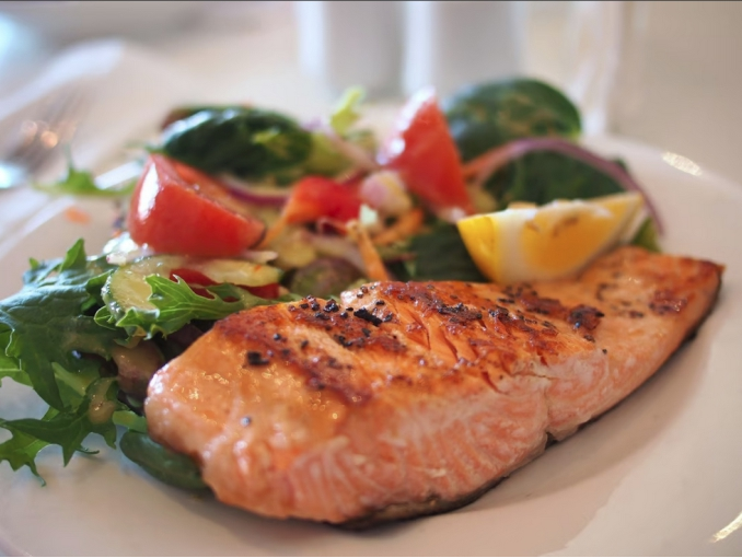 Auf einem weißen Teller liegt ein gebratenes Stück Lachs sowie ein gemischter Salat und ein Stück Zitrone.
