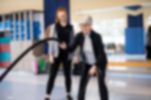 Eine Frau mit kurzen grauen Haaren trainiert an einem Seil unter Anleitung einer Trainerin