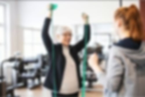 Eine Patientin trainiert im MTT und zieht mit ihren Armen Bänder in die Höhe, dabei wird sie von einer Therapeutin begleitet.