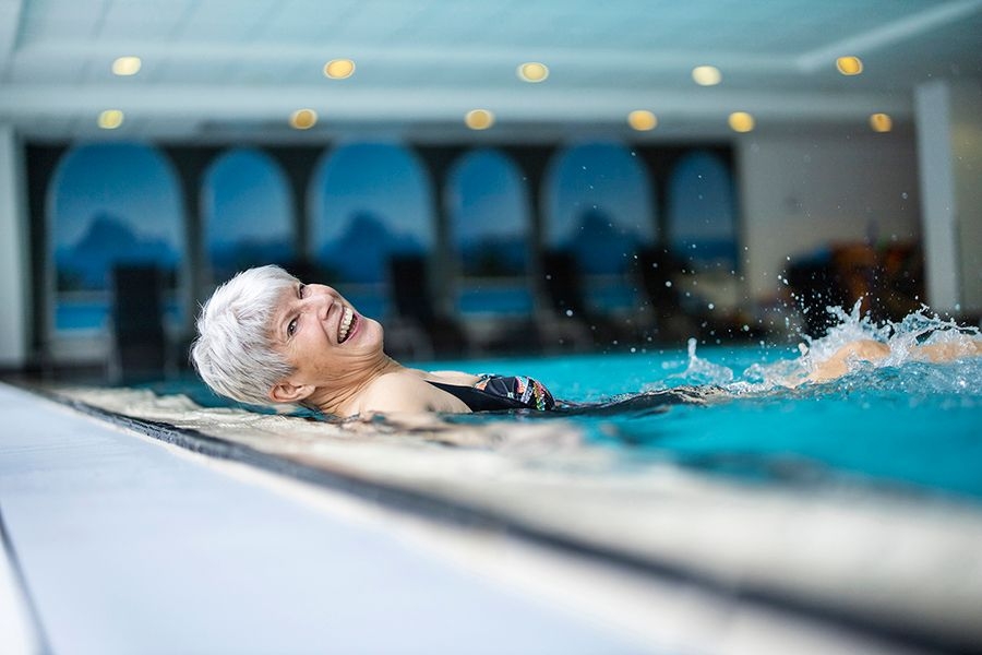 Eine Frau mittleren Alters mit kurzen grauen Haaren liegt im Schwimmbad im Wasser und lacht