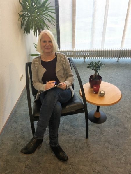 Die Psychologin der Klinik Solequelle sitz auf einem Stuhl neben einem Beistelltisch mit einer Blume darauf und lächelt.