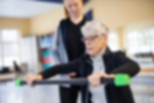 Eine Frau hält eine Trainingsstange in beiden Händen und wird von einer Trainerin angeleitet