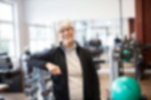 Eine Frau lehnt im Fitnessraum an einem Ständer mit Hanteln und lächelt in die Kamera.