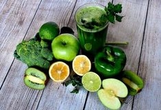 Ein Apfel, eine Paprika, eine Kiwi, eine Limette und eine Glas mit grünem Smoothie stehen auf einem Holztisch