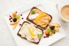 Auf einem weißen Teller liegen zwei Scheiben Brot mit Aufstrich und verschiebenen Obststücken. Daneben steht eine Tasse Kaffee.