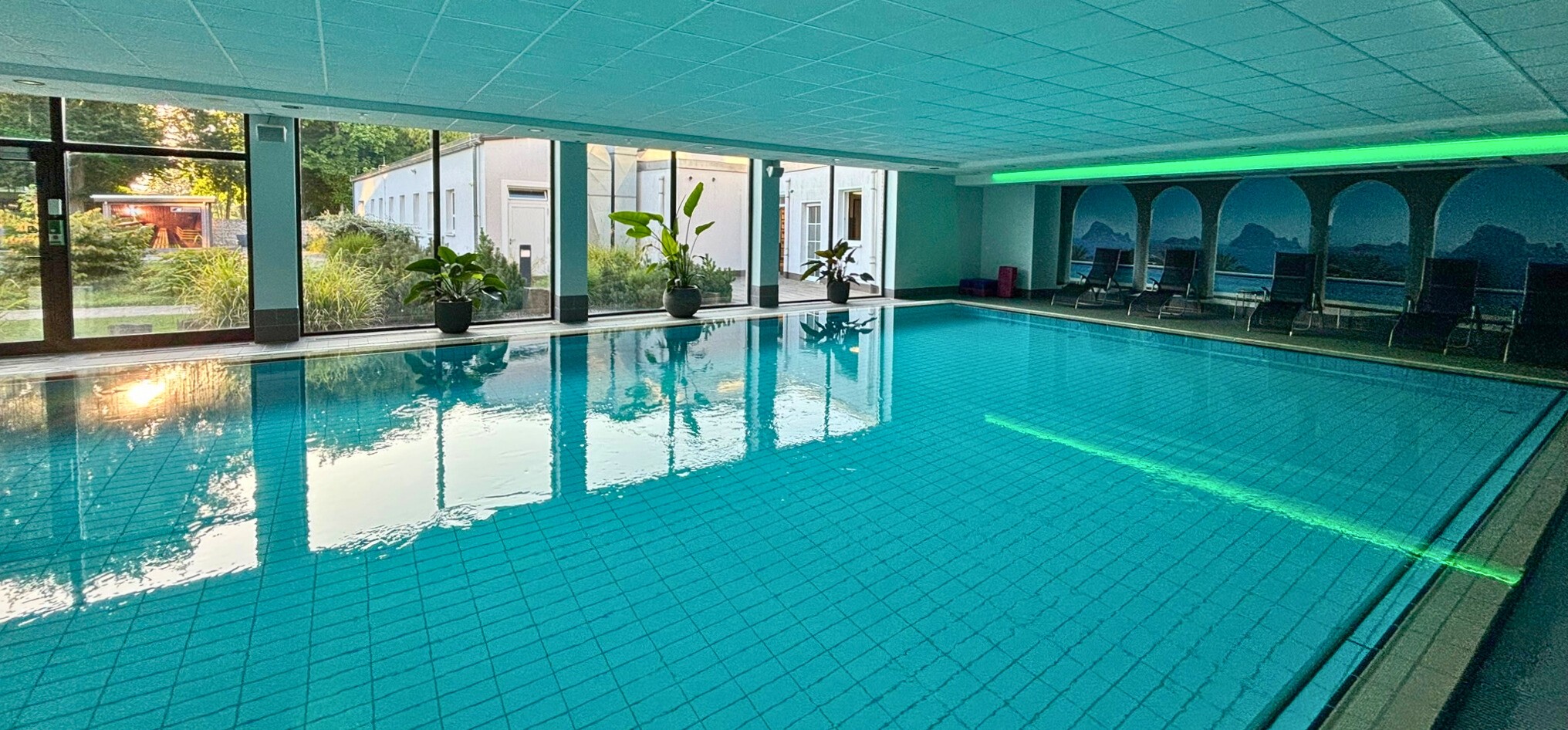 Blick auf unser hauseigenes Sole-Thermalschwimmbad. Am rechten Beckenrand stehen Stühle und Liegen zur Verfügung. Auf der linken Seite ermöglicht eine große Fensterfront den Blick auf unseren Klinikgarten.