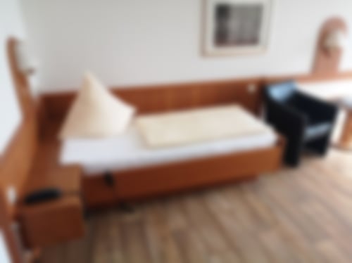 Ein Einzelzimmer mit Einzelbett und Nachttisch, TV, Schreibtisch, Sessel und einer großen Fensterfront zum eigenem Balkon.