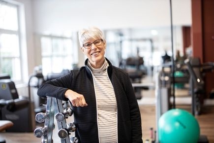 Eine Frau mittleren Alters mit kurzen grauen Haaren lehnt in einem Fitnessstudio an einem Hantelregal.