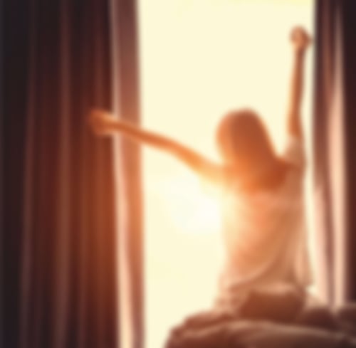 Eine Frau sitzt auf einem Bett und streckt sich. Im Hintergrund ist ein Fenster, durch das die Sonne scheint
