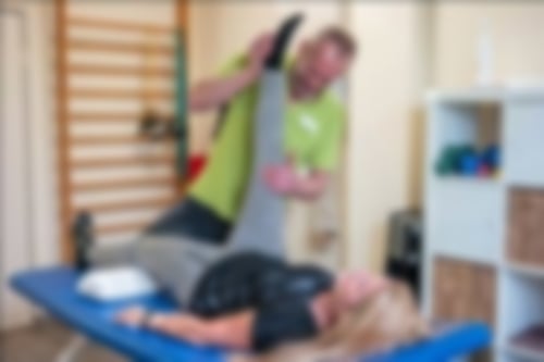 Eine Patientin liegt auf einer Liege in einem Therapieraum und führt mit Hilfe eines Therapeuten Übungen durch. Dabei streckt sie ihr Bein nach oben.