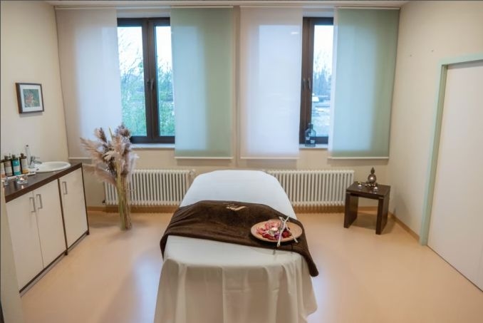 Eine Liege in einem unserer Wellnessräume. Auf der Liege liegt ein Handtuch und einer Dekoteller mit Blütenblättern und einer Kerze. Links steht ein Schraknk und Deko, rechts ein kleiner Beistelltisch mit einer Buddah-Statue.