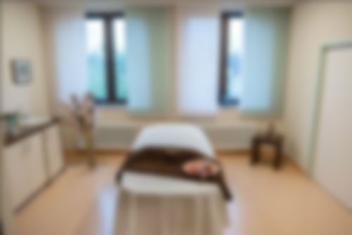 Eine Liege in einem unserer Wellnessräume. Auf der Liege liegt ein Handtuch und einer Dekoteller mit Blütenblättern und einer Kerze. Links steht ein Schraknk und Deko, rechts ein kleiner Beistelltisch mit einer Buddah-Statue.