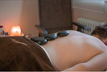 Ein Mann liegt auf einer Liege und entspannt bei einer Hot-Stone Massage. Die warmen Steine liegen auf seinem Rücken. Im Hintergrund leuchtet ein Salzkristall und sorgt für Gemütlichkeit.