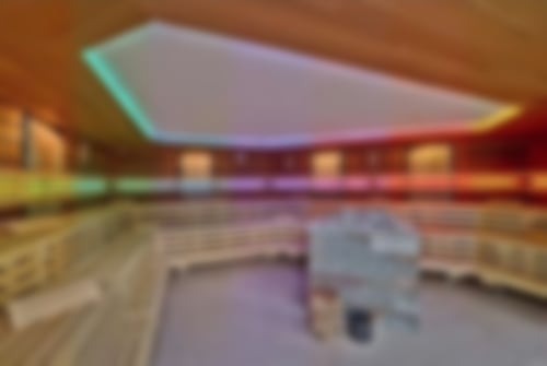 Innenansicht unserer Anröchter Grünsandstein-Sauna. Am Rand befinden sich die Bänke, darüber bunte Lichter. In der Mitte steht der Ofen mit den Steinen, davor ein Holzblock und ein Eimer.