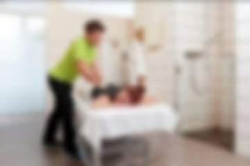Eine Frau liegt auf dem Bauch auf einer Liege und erhält eine Moor-Rückenpackung von einer Therapeutin. Rechts im Hintergrund befindet sich eine Dusche.