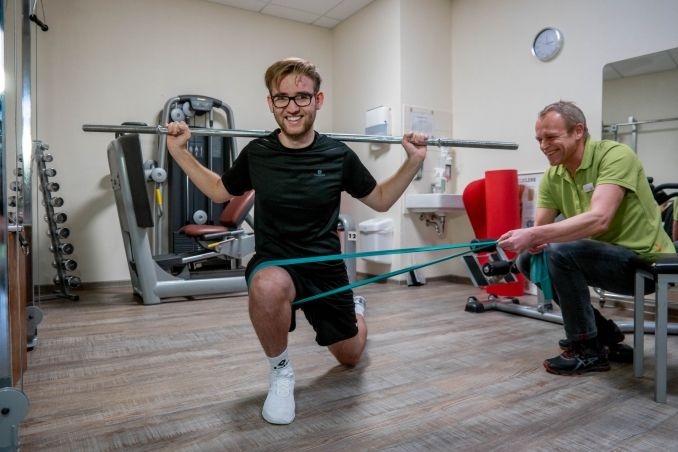 Ein Mann kniet auf dem Boden und hebt hinter seinem Rücken eine Stange über seine Schultern. Daneben ist ein Therapeut, der an einem elastischen Band zieht, welches um das Bein das Mannes gewickelt ist.