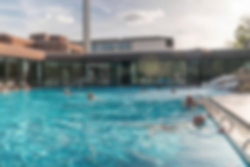 Unser großzügiger Außenbereich. Im Pool schwimmen mehrere Personen, manche entspannen am Beckenrand. Im Hintergrund ist das Gebäude zu sehen.