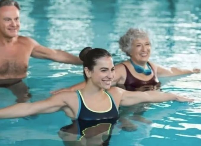 Ein Mann und zwei Frauen befinden sich im Schwimmbecken und führen Übungen durch. Dabei strecken sie ihre Arme seitlich von Körper aus.