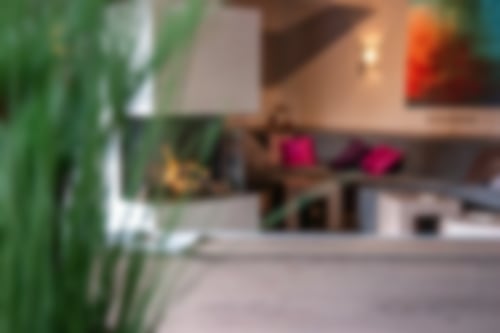 Unsere gemütliche Kamin-Lounge. In der Mitte steht ein großer Kamin, in dem hinter einer Glaswand Feuer brennt. Auf der rechten Seite lädt ein Sofa mit Kissen zum Entspannen ein. Davor stehen kleine Tische.