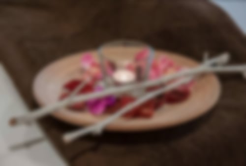 Auf einem braunen Handtuck steht ein Dekoteller mit Blütenblättern, zwei Ästen und einem Glas, in dem ein Teelicht brennt,