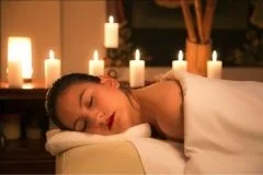 Eine Frau liegt auf dem Bauch auf einer Liege, mit dem Kopf zur Seite. Im Hintergrund sorgen brennende Kerzen für Entspannung.