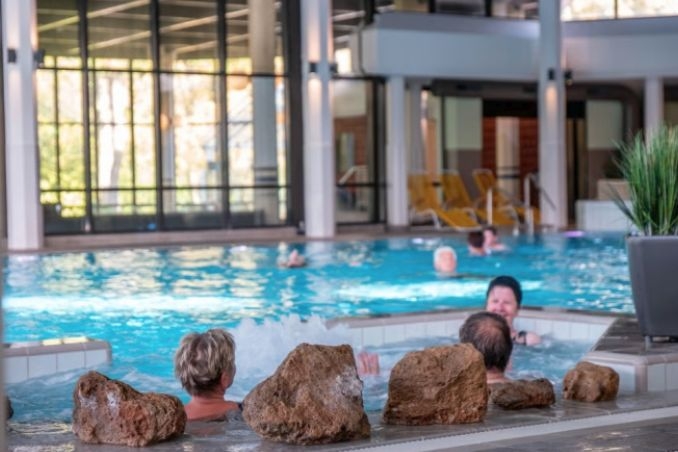 Unsere Gäste schwimmen im großzügigen Innenbecken. Im Vordergrund befindet sich ein Whirlpool, in dem mehrere Personen sitzen und entspannen. Im Hintergrund stehen Liegen bereit.