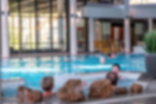 Unsere Gäste schwimmen im großzügigen Innenbecken. Im Vordergrund befindet sich ein Whirlpool, in dem mehrere Personen sitzen und entspannen. Im Hintergrund stehen Liegen bereit.