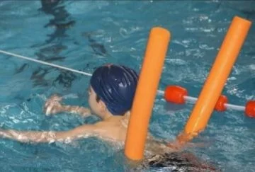 Ein kleiner Junge mit Badekappe schwimmt mit Hilfe einer Schwimmnudel durchs Wasser.
