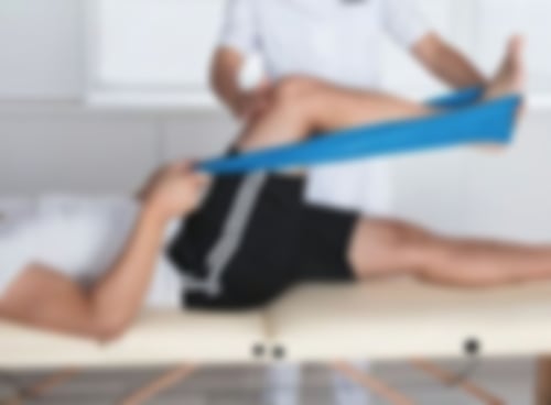 Ein Mann liegt auf einer liege und macht Beinübungen mit einem blauen Theraband, welches um seinen Fuß gebunden ist und er mit den Händen zieht. Ein Therapeut steht neben ihm und unterstützt bei den Übungen.