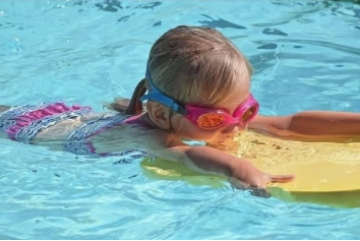 Ein kleines Mädchen mit Taucherbrille paddelt mit einem Schwimmbrett durch das Wasser.