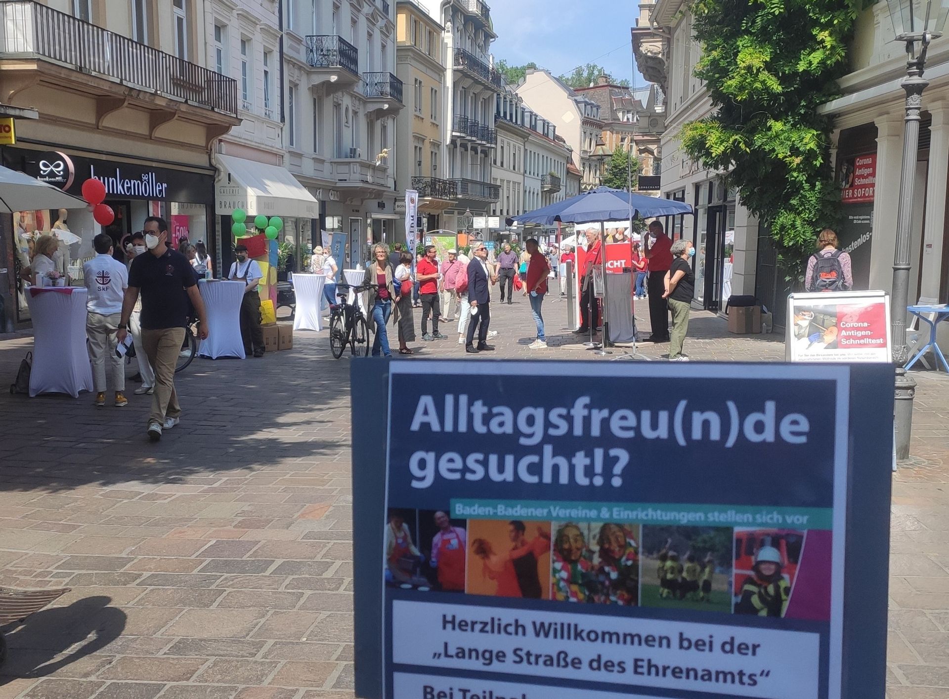 Fußgängerzone an einem sonnigen Tag. Im Vordergrund ein Schild mit der Aufschrift Alltagsfreunde gesucht?!