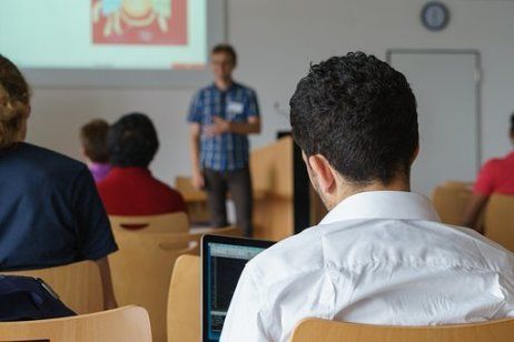 im Vordergrund Menschen, die in einem Vortrag sitzen. Im Hintergrund ein stehende Person vor einer Präsentation