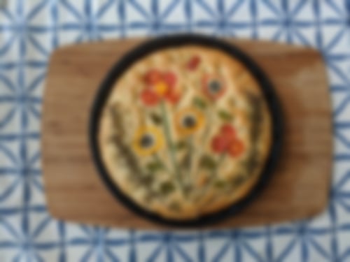 Hausgemachte Focaccia mit kunstvoll arrangierten Gemüsedekorationen aus Tomaten, Paprika, Oliven und Kräutern wie Rosmarin und Schnittlauch.