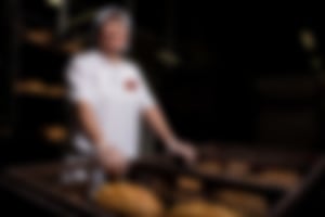 Mitarbeiterin in Essmann’s Backstube-Uniform und Haube steht lächelnd an Transportkisten voller frisch gebackener Brote in der Backstube.