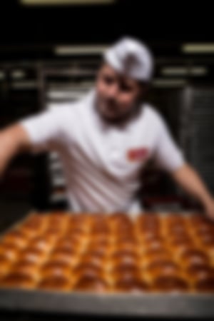 Bäcker in Essmann’s Backstube-Uniform hält ein großes Blech mit frisch gebackenen, goldbraunen Rosinenbrötchen in der Backstube.