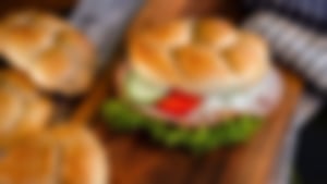 Frisch belegtes Sesambrötchen mit Salat, Tomaten, Gurken, Krautsalat und Wurst auf einem Holzbrett, umgeben von weiteren frischen Brötchen.