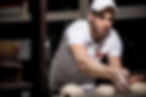 Bäcker in weißer Essmann’s Backstube-Uniform präsentiert stolz ein großes Blech voller frisch gebackener, goldbrauner Rosinenbrötchen in der Backstube.